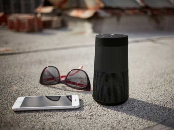 The Bose SoundLink Revolve II on the sidewalk with a pair of glasses and smartphone.