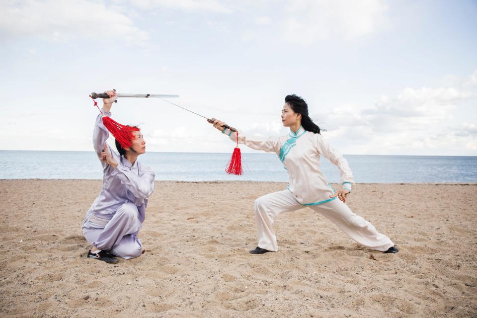Two tai chi practioners performing a routine with swords in traditional uniforms on the beach.