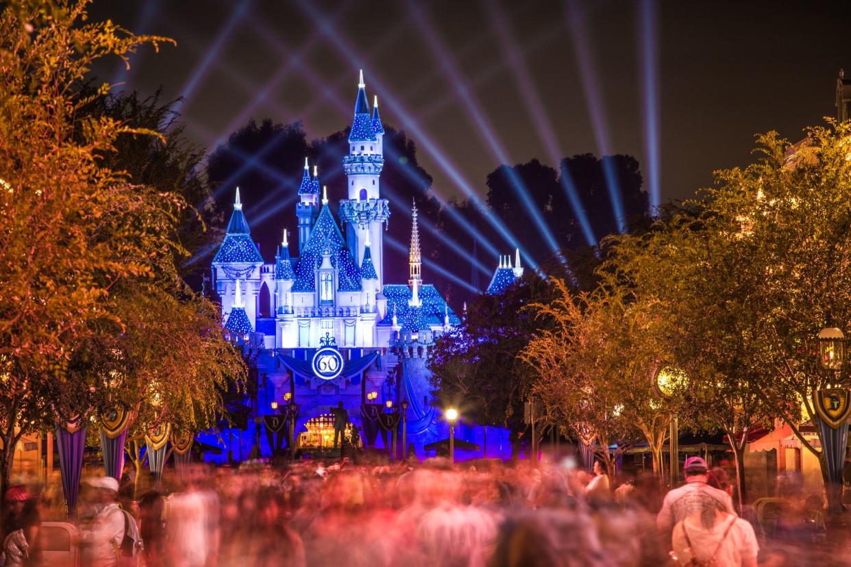 60th anniversary celebration in Disneyland, Anaheim, California, during evening, blue lit castle in the background with blurred pink-brown lit people in the foreground, dramatic lights surrounding castle