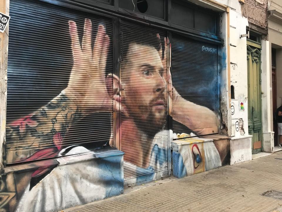 street mural on black wall of lionel messi in argentina jersey with hands up to his ears