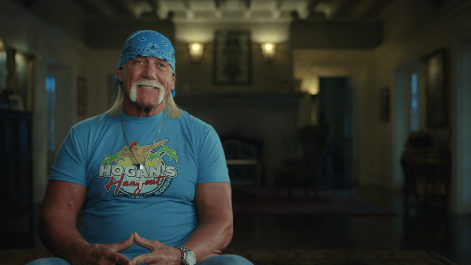 A smiling older man with tan light-toned skin and a bushy blond mustache/fu manchu, sitting in a nice living room-esque setting.