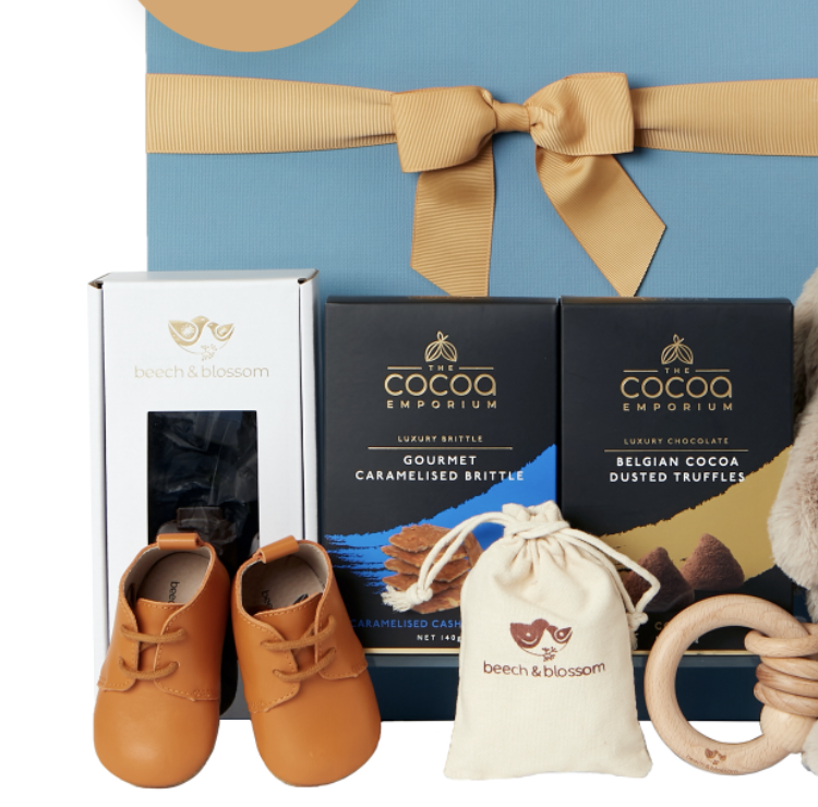 The Gourmet Pantry Sweet Baby Bundle Hamper, $129, is shown with cocoa products packaged in black boxes, a white box of chocolates, tan soft baby shoes, wooden rings and a drawstring bag from Beech & Blossom 