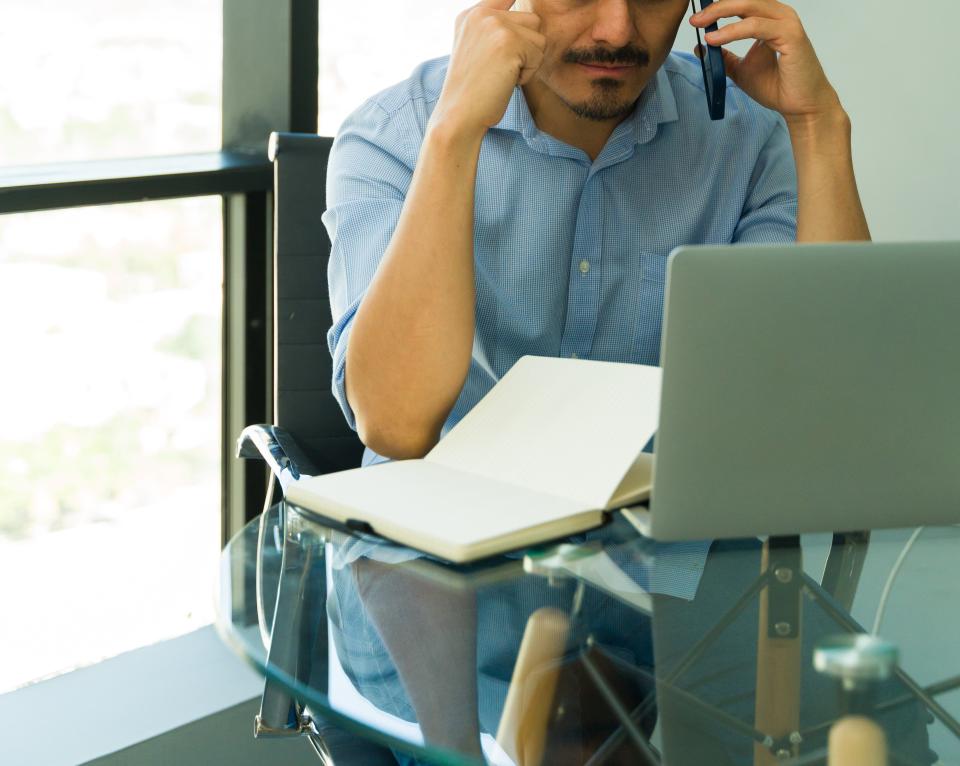 Man in a blue shirt sits at a glass table with an open notebook and laptop, talking on the phone and touching his temple