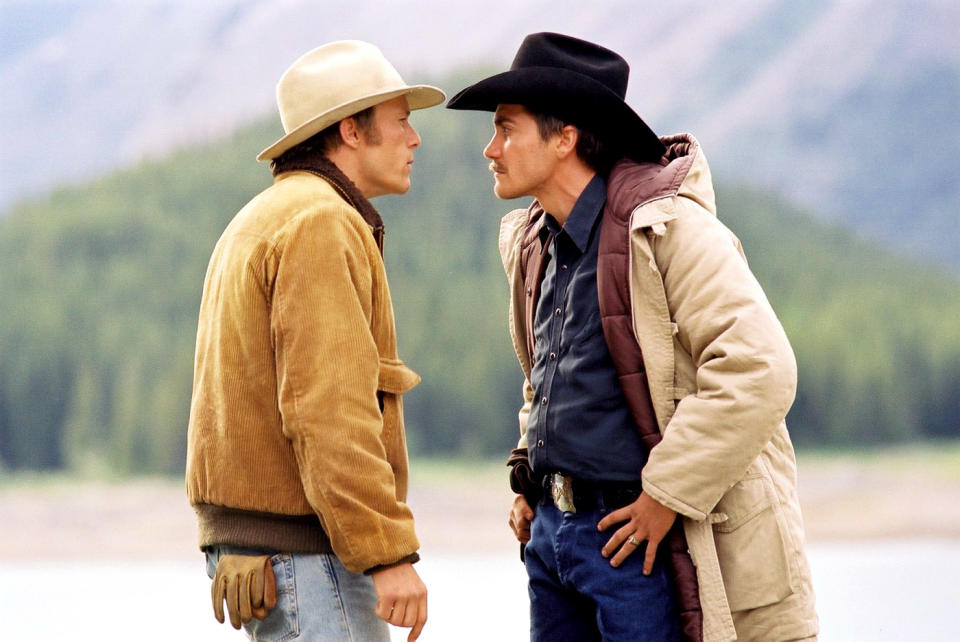 Heath Ledger and Jake Gyllenhaal stand facing each other in a tense scene from "Brokeback Mountain," both wearing cowboy hats and layered jackets. Mountains are visible in the background