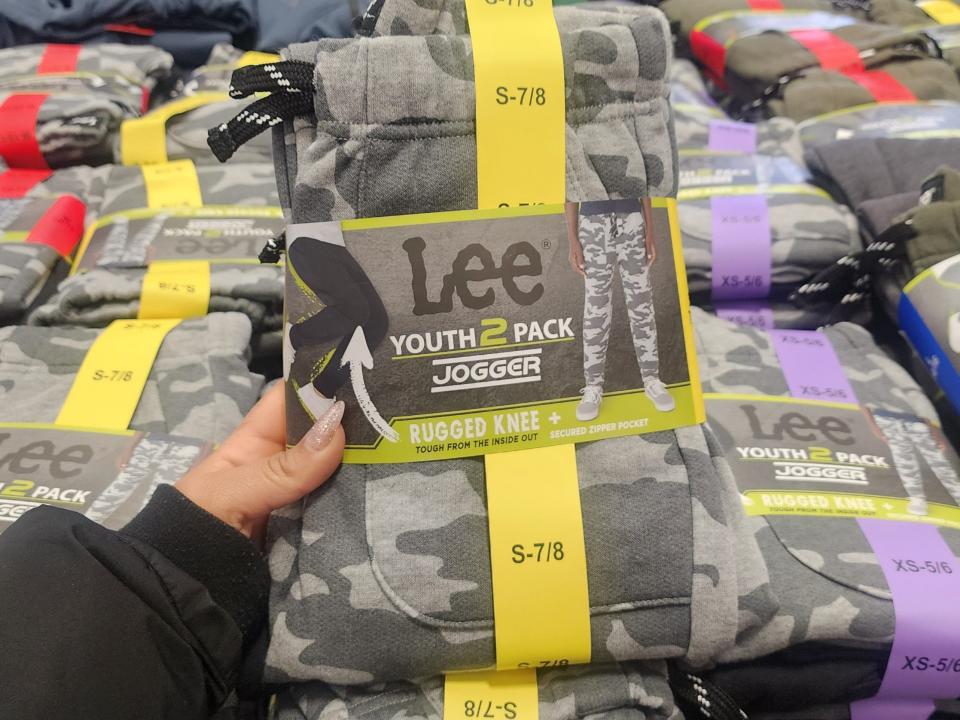 Hand holding dark and light gray camouflage pants. A label on the pants say "Lee Youth 2 Pack Jogger"