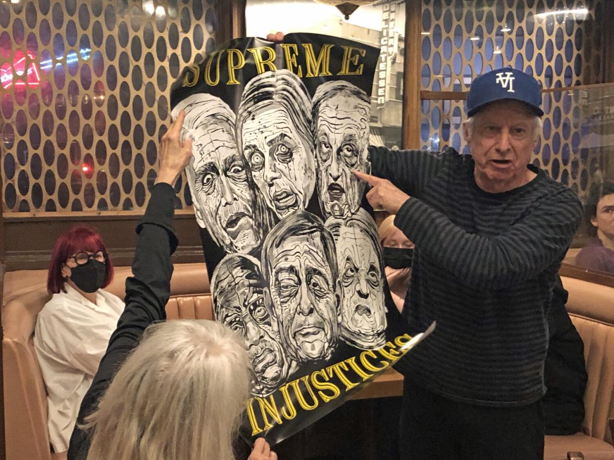 Artist Robbie Conal holds up a poster titled "Supreme Injustices," showing six members of the U.S. Supreme Court