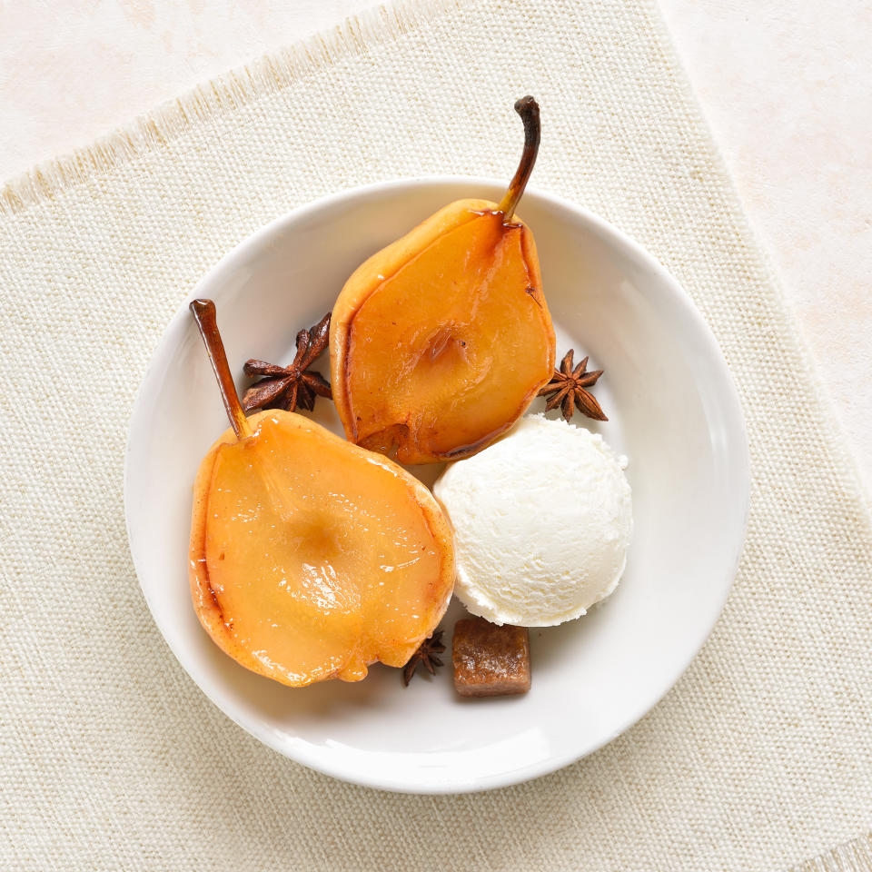 A white bowl with two poached pear halves, a scoop of vanilla ice cream, and star anise. The dessert is placed on a beige fabric surface