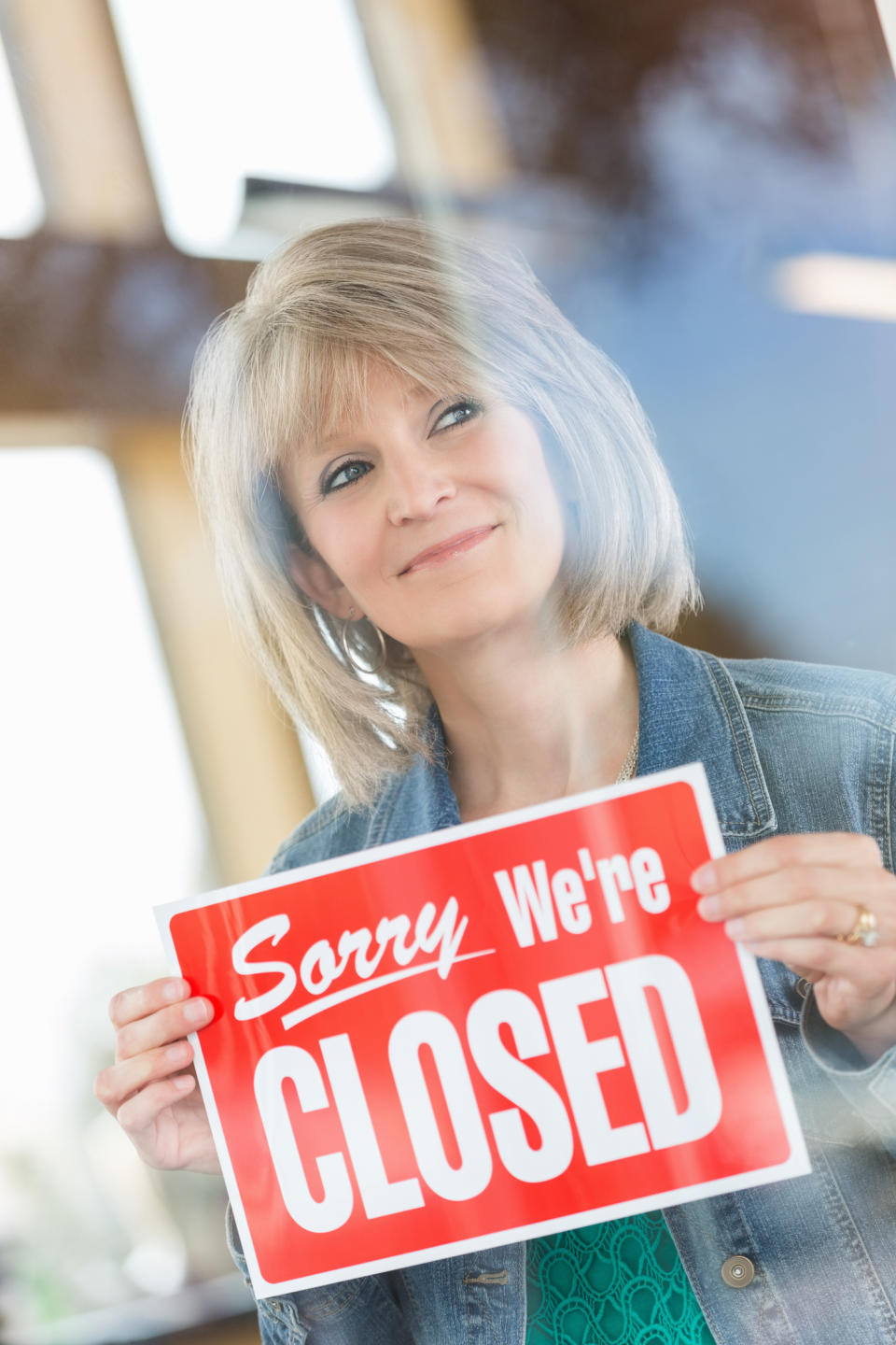 Woman holding a 'Sorry We're Closed' sign, viewed through a glass pane