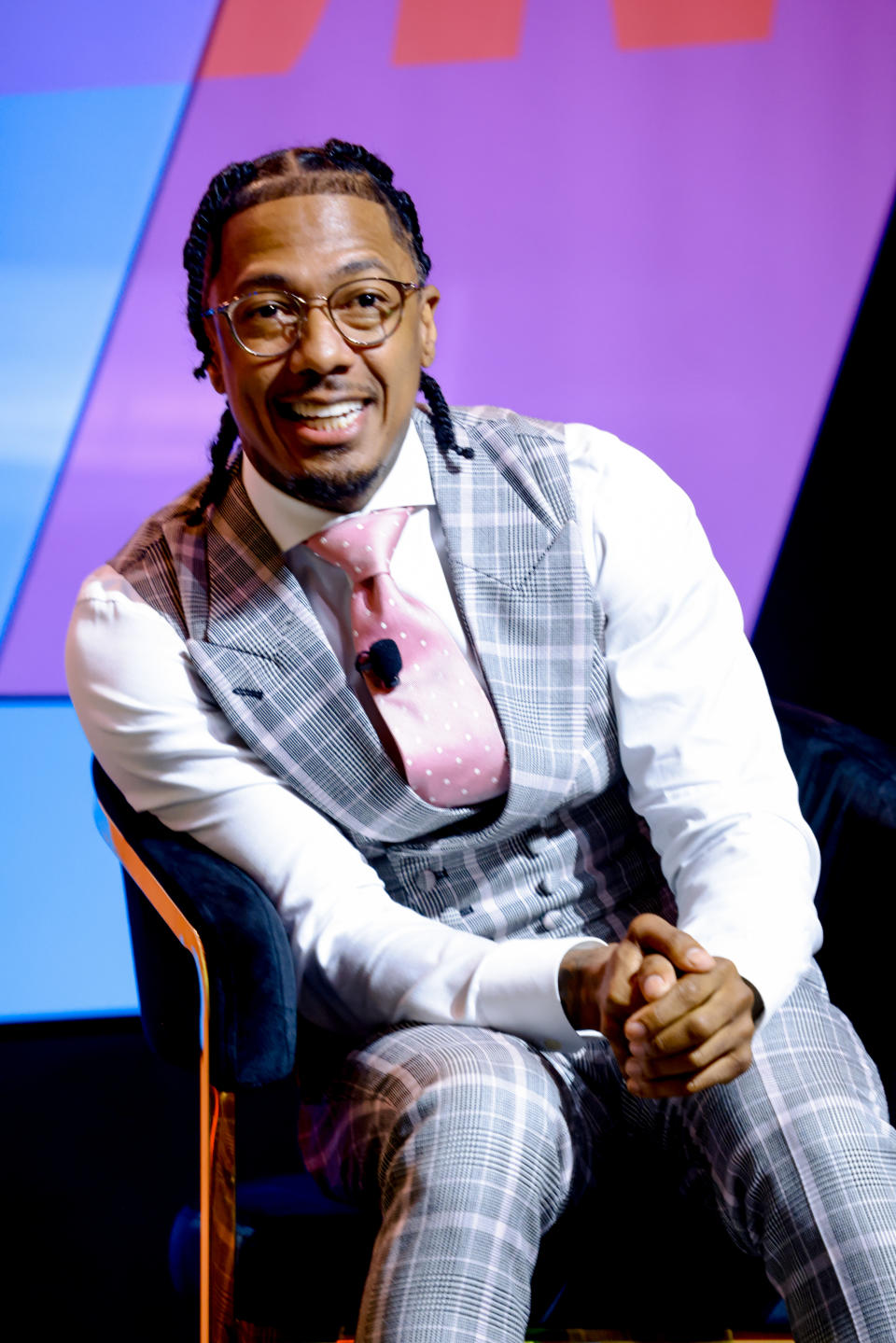 Nick Cannon sits on a chair wearing a plaid suit, white shirt, and pink polka dot tie, smiling at an event