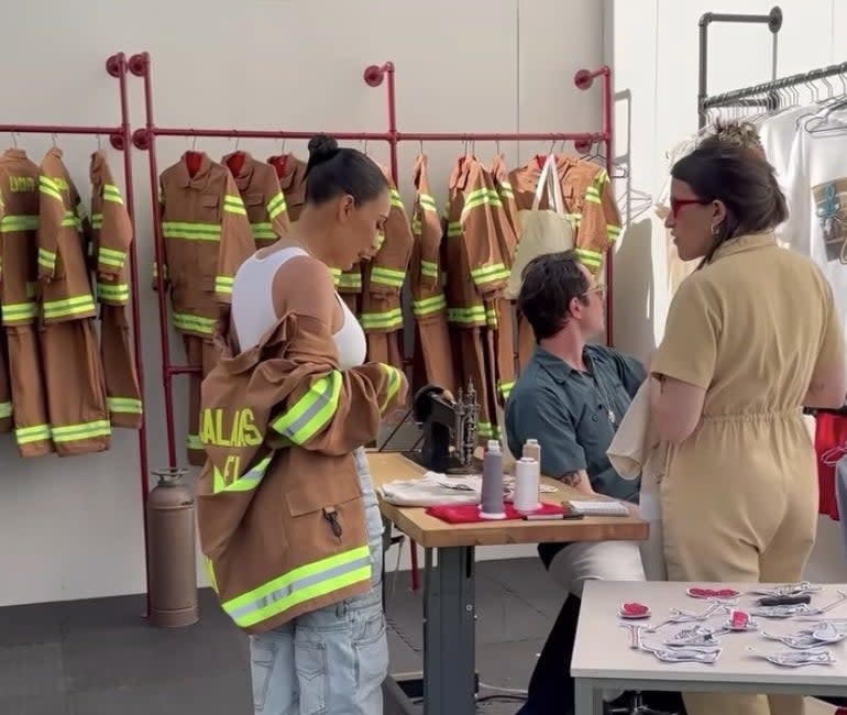 Kim stands in front of a table with embroidery equipment and t-shirts and jackets are hung up on a wall behind her