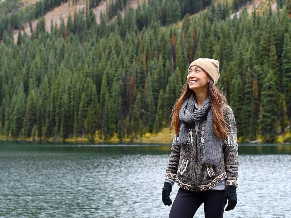 The writer dressed for winter standing in front of a lake that is surrounded by evergreen trees