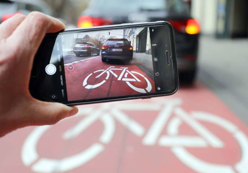 A picture of a car parked on a cycle lane can be seen on a smartphone. A growing number of parking offences are being reported by private individuals across Germany. Oliver Berg/dpa