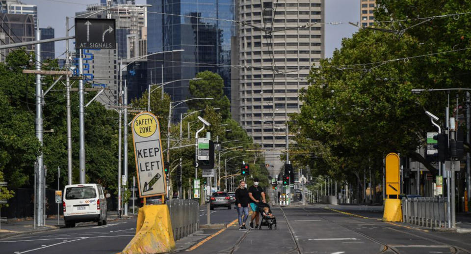 Two people cross a Melbourne street with face masks, pushing a pram with a child in it.