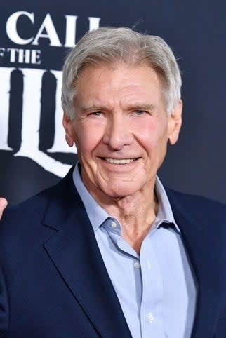 Harrison Ford waving at a premiere, wearing a suit jacket over a shirt, backdrop features 'The Call of the Wild'