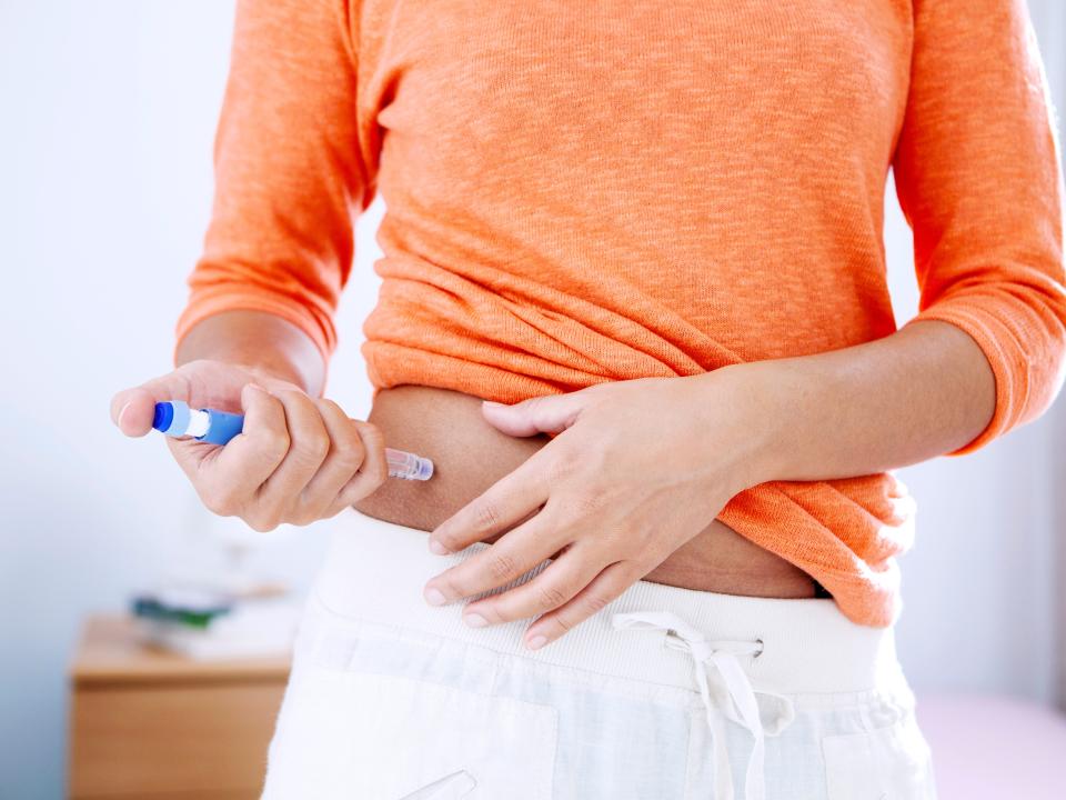 A woman in an orange shirt using an injection pen on the right side of her stomach.