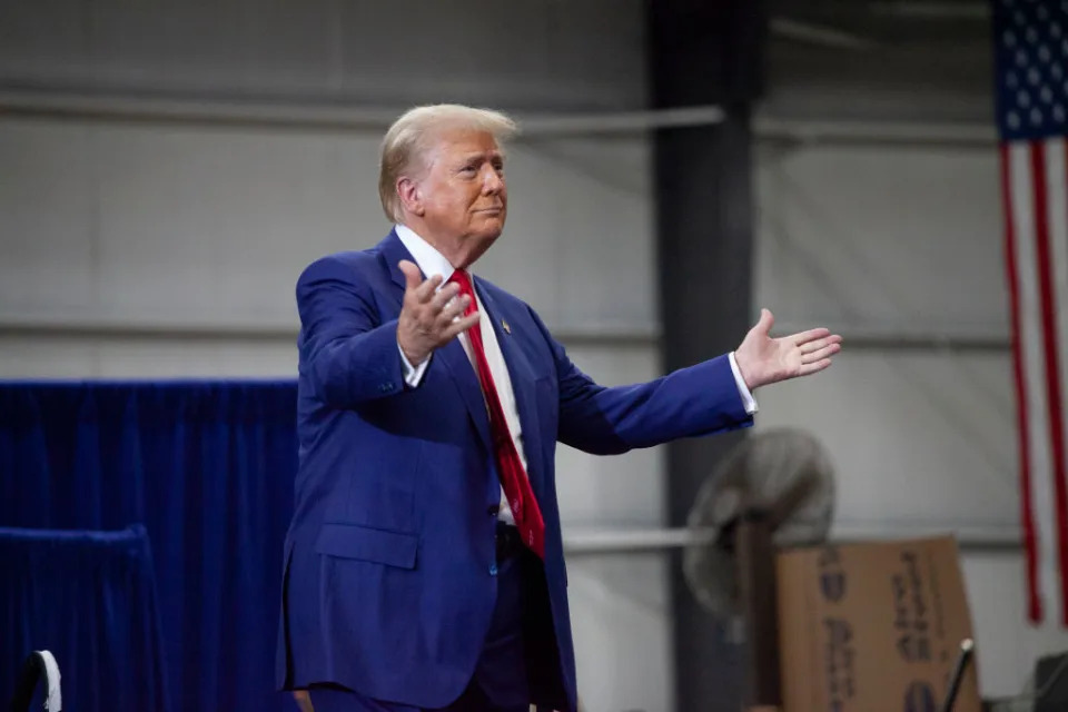 Donald Trump gestures with open arms at an indoor event, standing near a U.S. flag