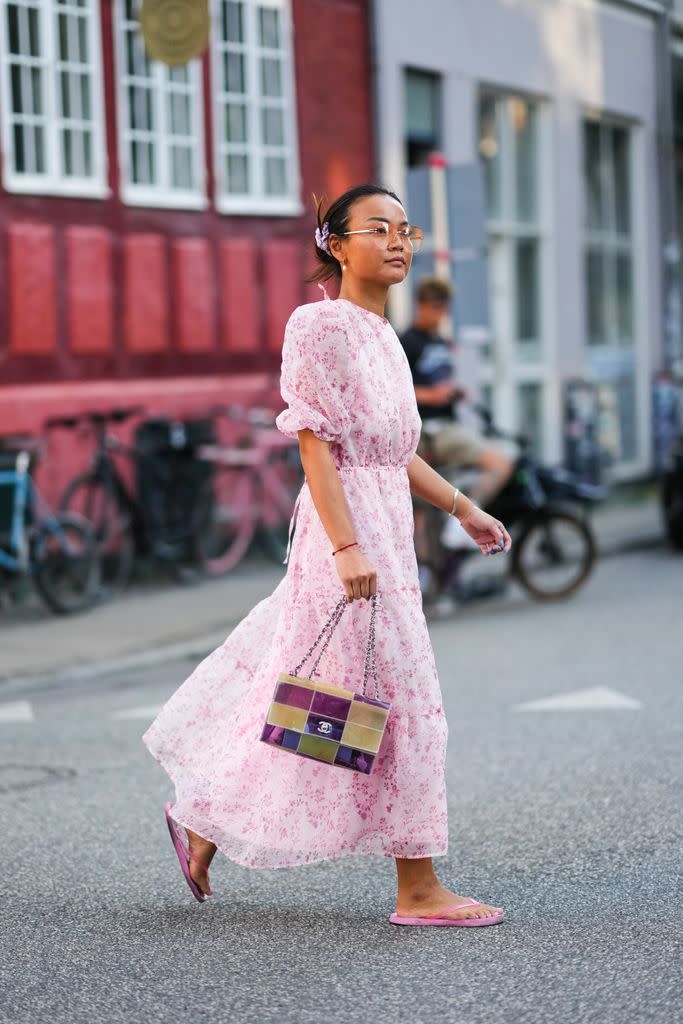 COPENHAGUE, DINAMARCA - 10 DE AGOSTO: Una invitada lleva gafas de sol naranjas, pendientes dorados, un vestido midi blanco con estampado de flores rosa pálido / hombros abullonados / mangas cortas / volantes, una pulsera roja, un bolso de plástico brillante amarillo y morado con estampado de cuadros de Chanel, chanclas rosas , fuera de Baum und Pferdgarten, durante la Semana de la Moda de Copenhague Primavera/Verano 2023, el 10 de agosto de 2022 en Copenhague, Dinamarca. (Foto de Edward Berthelot/Getty Images)