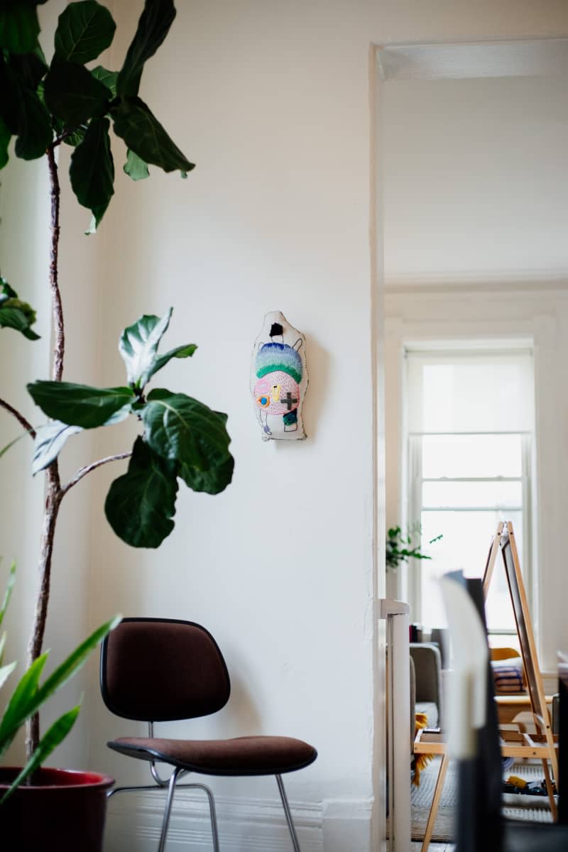 Embroidered pillow hung on wall above stool in light filled apartment.