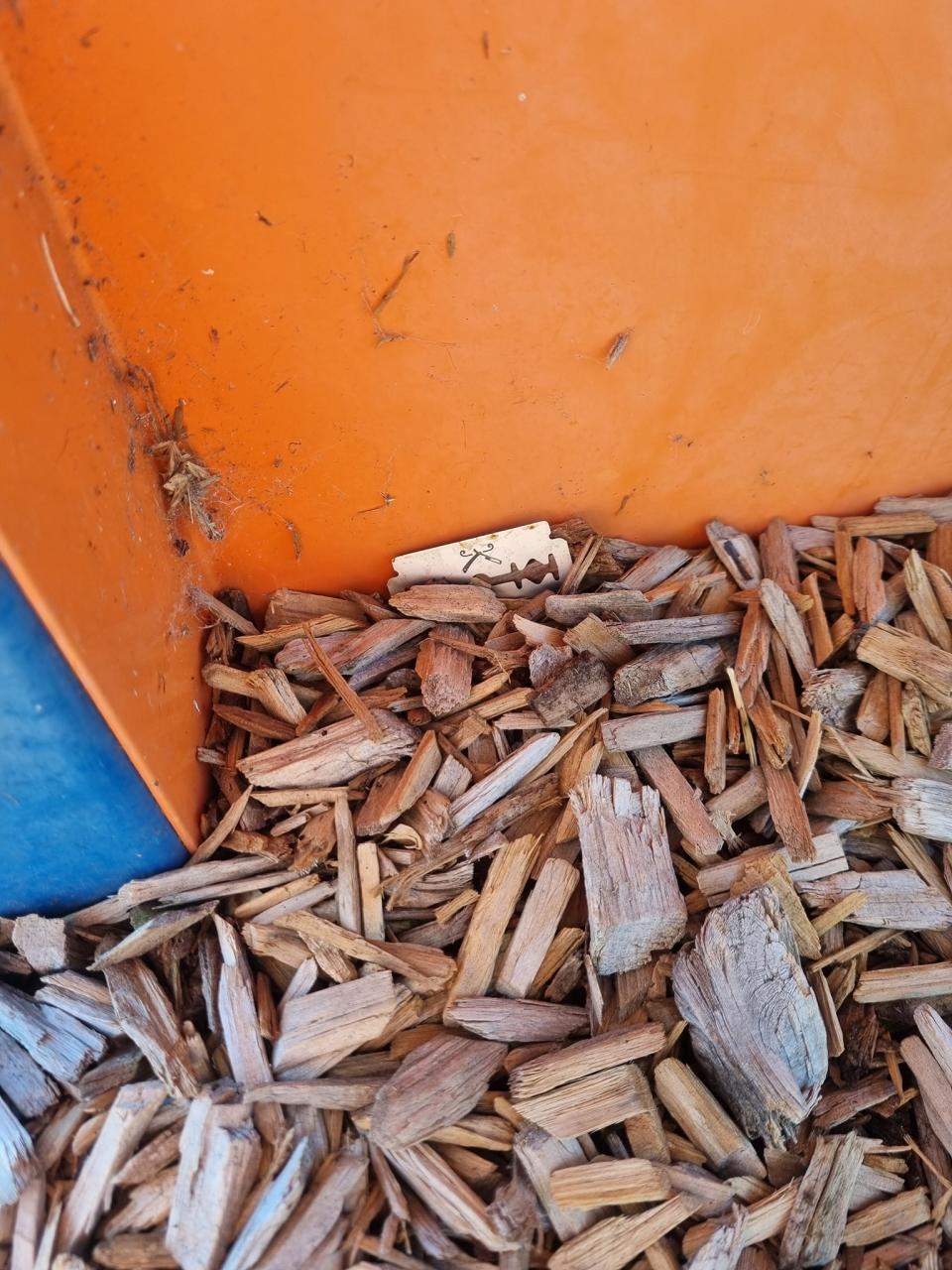 The side of an orange piece of playground equipment with brown chip bark, a silver blade is seen near the edge of the orange edge of playground equipment.