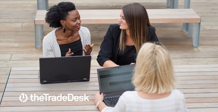 Three women sitting at a picnic table with laptops. The Trade Desk logo is in the lower left-hand corner..