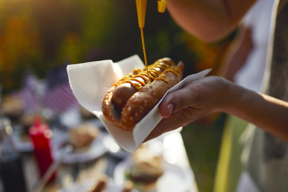 Close-up of a person adding mustard to a hot dog at an outdoor gathering, with blurred background of food and decor