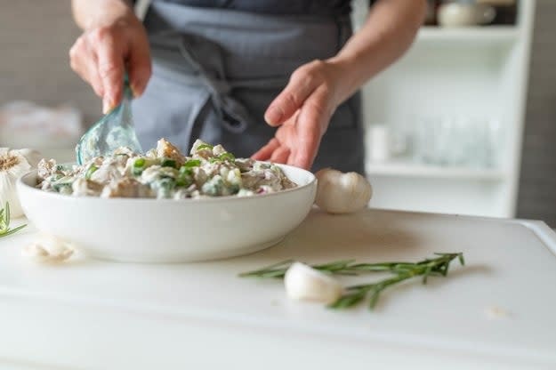 Person adding a final ingredient to a salad in a white bowl on a kitchen counter, with fresh herbs and garlic cloves around. Hands visible, face obscured