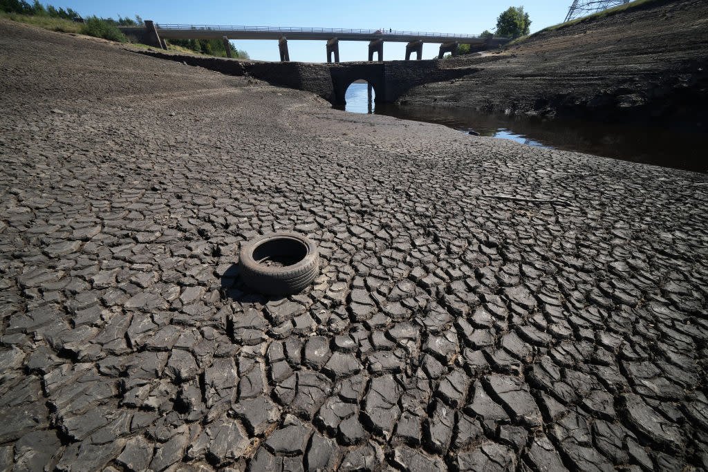 A rubber tire sits on the dry, cracked earth of a reservoir floor, with a two-tier bridge in the distance. 