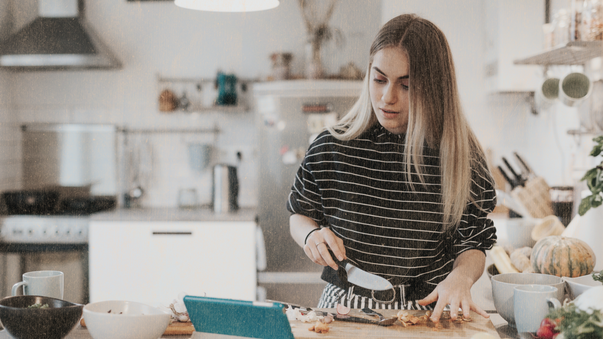 photo of a person standing in a home kitchen at a counter with a cutting board with food on it in front of them. There's a knife in their hand and they're looking at a tablet on the counter.