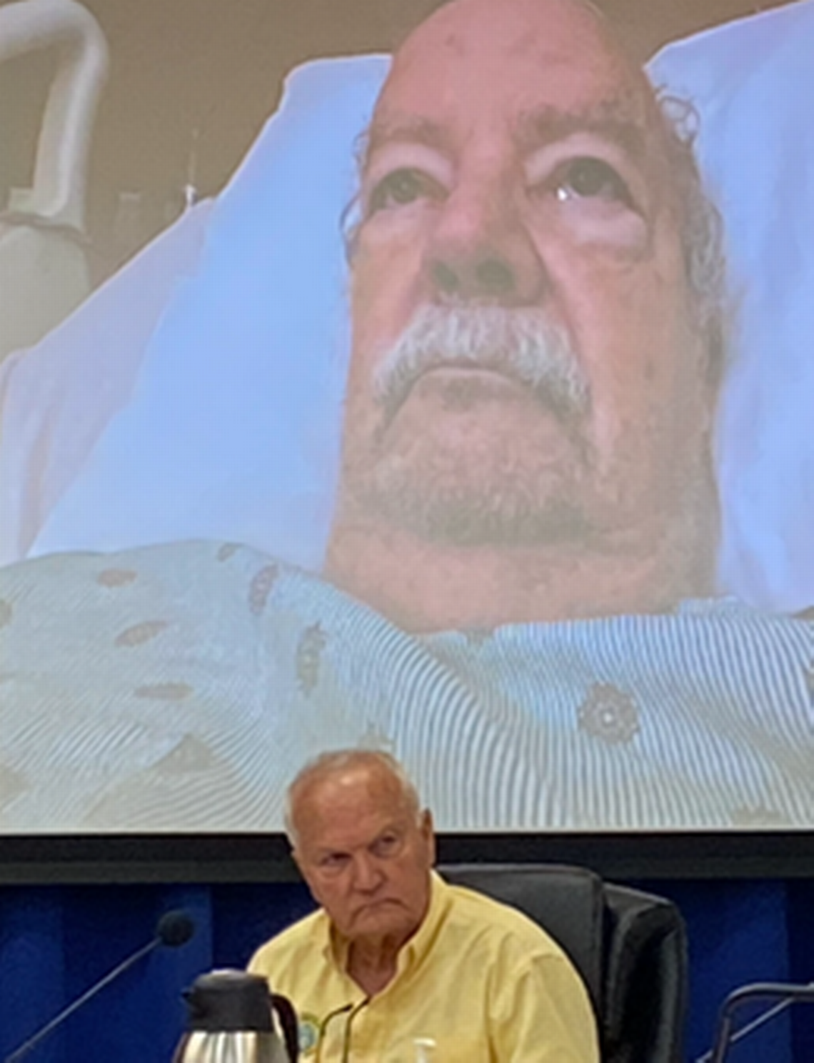 Islamorada Mayor Pete Bacheler appears on screen from his hospital bed during a Village Council meeting Thursday, Aug. 25, 2022. Sitting at the dais underneath the screen is Village Counciman Buddy Pinder. Details leading up to a vote taken during that meeting are under Monroe County State Attorney’s Office investigation.