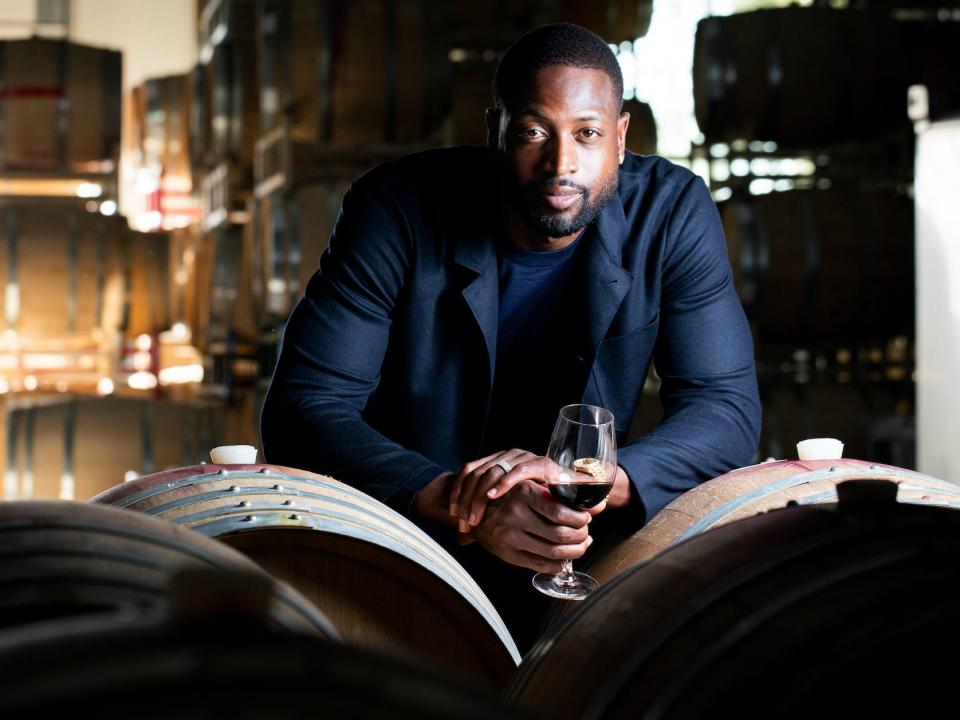 Dwyane Wade leans against barrels while holding a glass of wine.