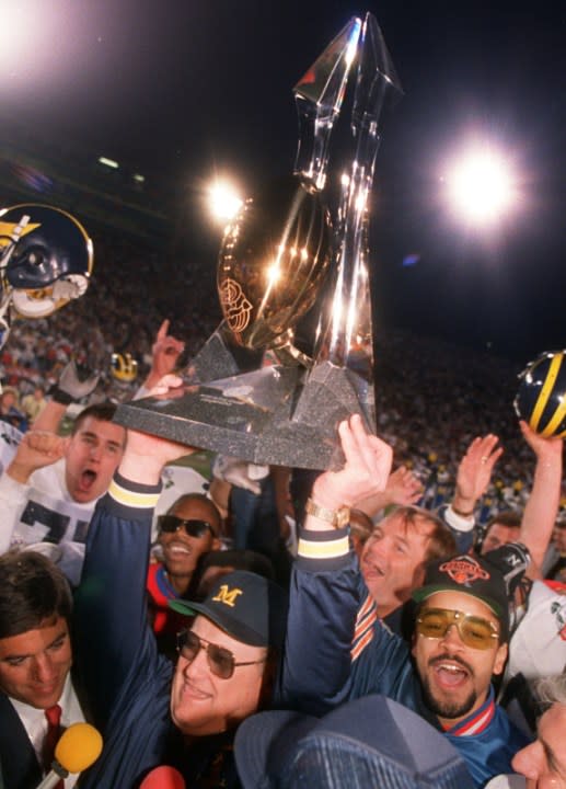 Surrounded by a horde of players and fans, Bo Schembechler holds the Rose Bowl Trophy above his head with both hands.