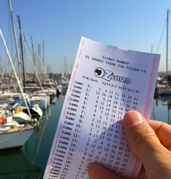 A man is seen holding a Lotto ticket in his hand with the ocean and boats floating in the background. Source: The Lott
