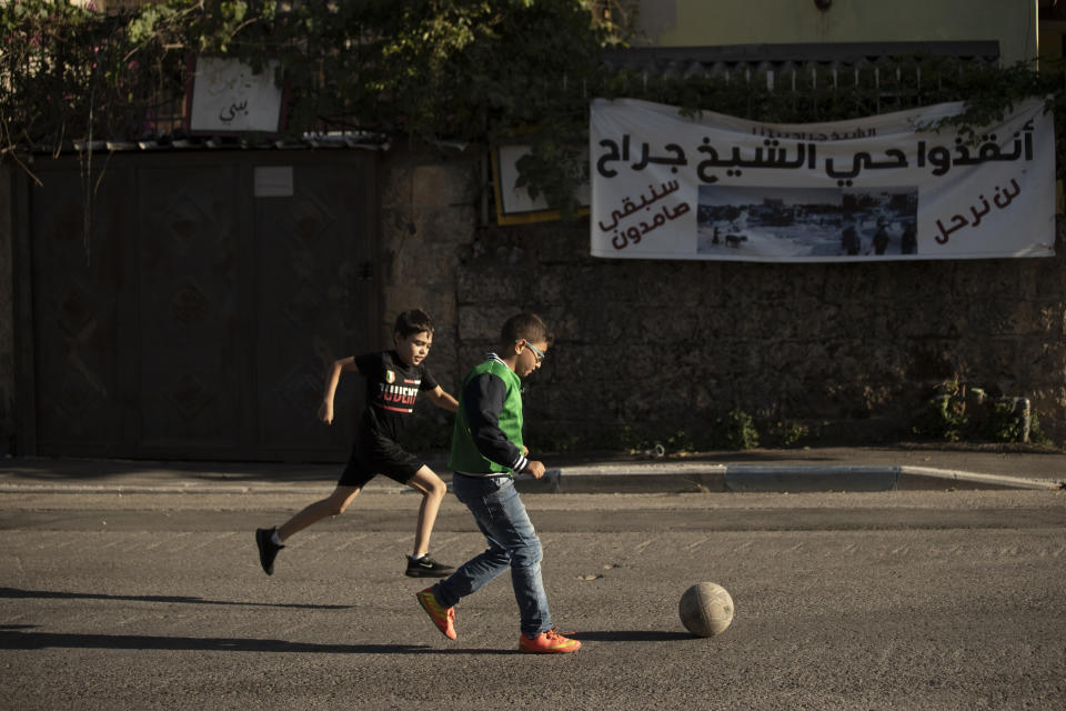 FILE - In this May 27, 2021, file, photo, Palestinian boys play soccer on their street in the Sheikh Jarrah neighborhood of east Jerusalem, where dozens of Palestinian families are facing imminent eviction from their homes by Israeli settlers. The banner behind them shows a vintage photo of their neighborhood, left, and a current image of a heavy security presence. Arabic reads: "Sheikh Jarrah neighborhood - we will not leave - we will remain steadfast." (AP Photo/Maya Alleruzzo, File)