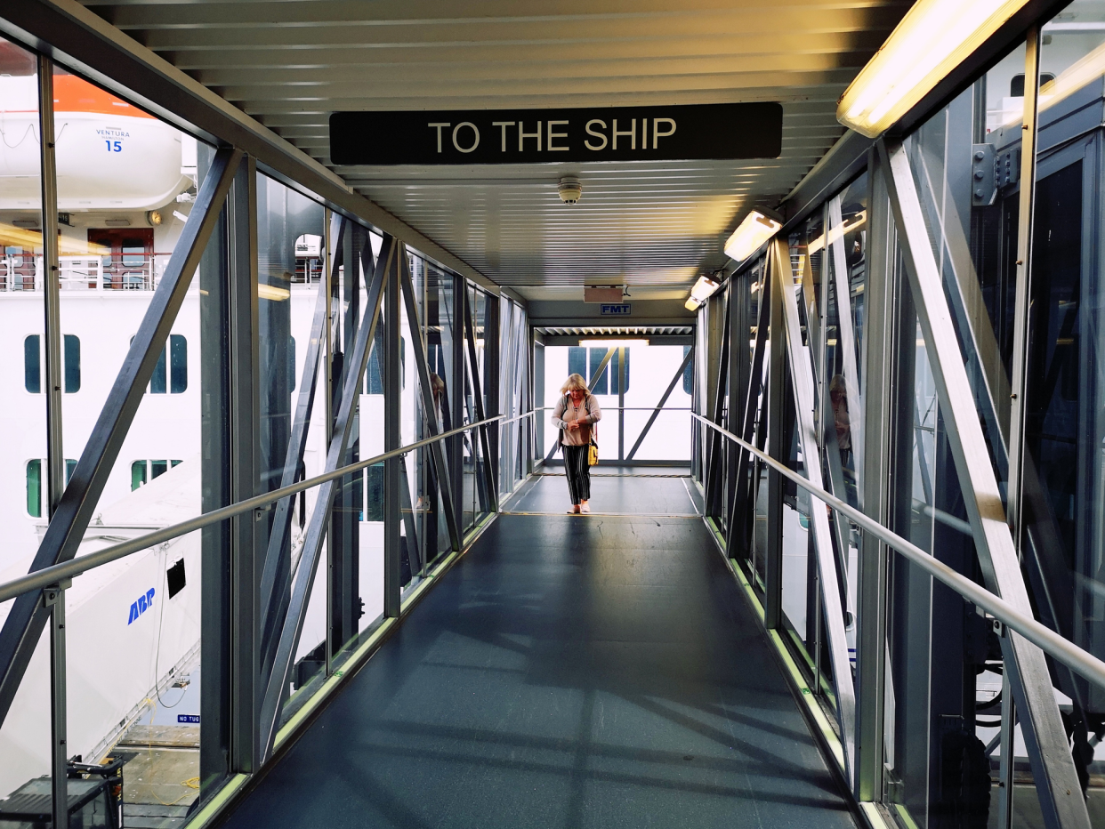 Female passenger checking watch as she goes through the boarding ramp of a cruise ship, empty ramp in the foreground with woman in the back of it, another cruise ship is dock in the background