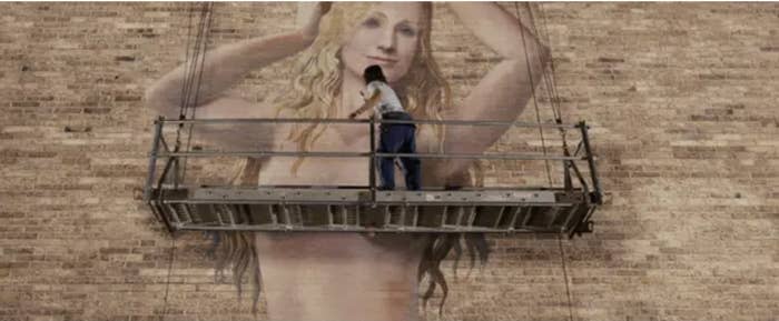 A person is working on a suspended platform, painting a large mural of a nude blonde woman on the side of a brick building