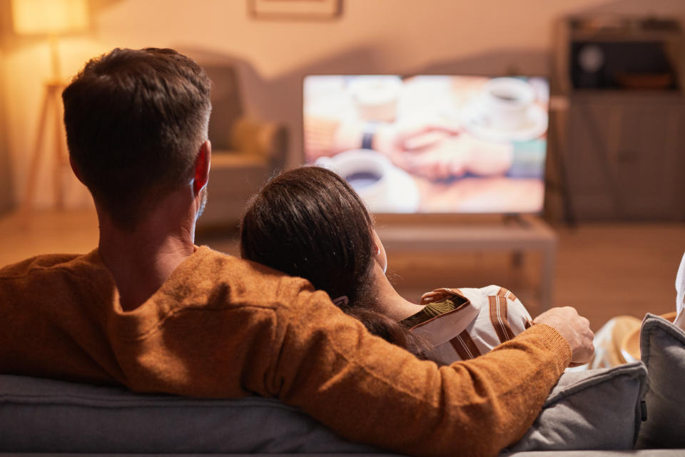 Back view of adult couple watching TV at home while sitting on sofa lit by warm cozy light, copy space