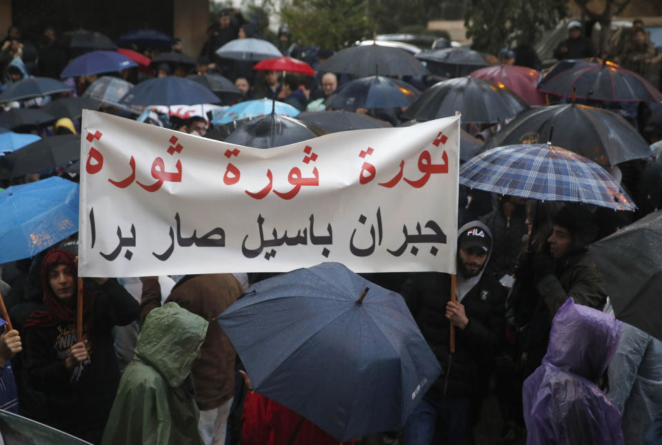 FILE - In this Sunday, Jan. 5, 2020 file photo, anti-government protesters hide under their umbrellas from the heavy rain, as they hold an Arabic banner that reads: "Revolution, Lebanese foreign minister Gebran Bassil is out," during a protest outside the residence of Lebanon's Prime Minister-Designate Hassan Diab, in Beirut, Lebanon. Lebanese are campaigning against the participation of their acting foreign minister in the prestigious World Economic Forum in Davos, saying Bassil, who was the target of popular anger in the ongoing nationwide protests, should not represent them on the international stage. (AP Photo/Hussein Malla, File)