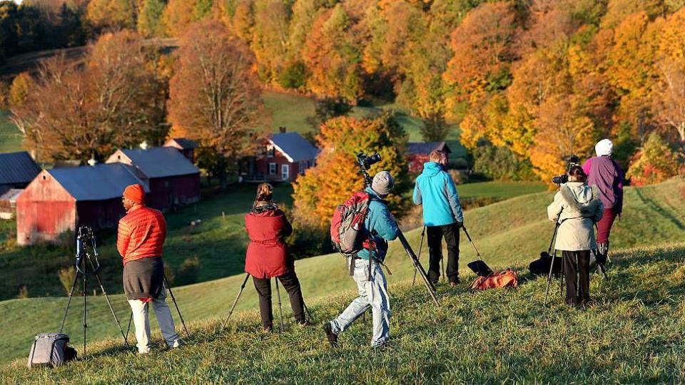 Turistas con sus cámaras y trípodes frente a una granja con árboles de colores otoñales  / Foto: Getty Image 