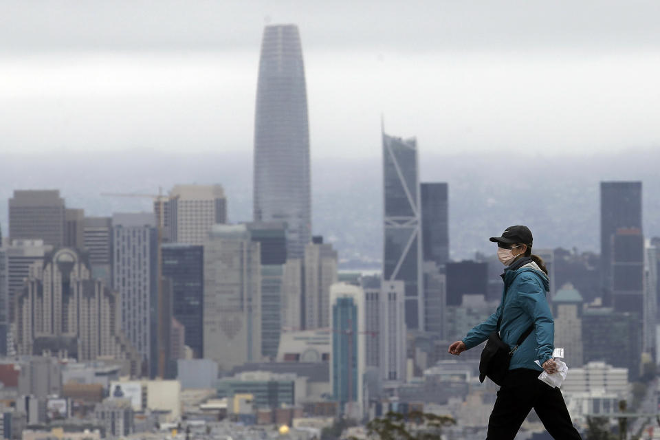 A woman wears a mask during the coronavirus outbreak while crossing a street in front of the skyline in San Francisco, Saturday, April 4, 2020. (AP Photo/Jeff Chiu)