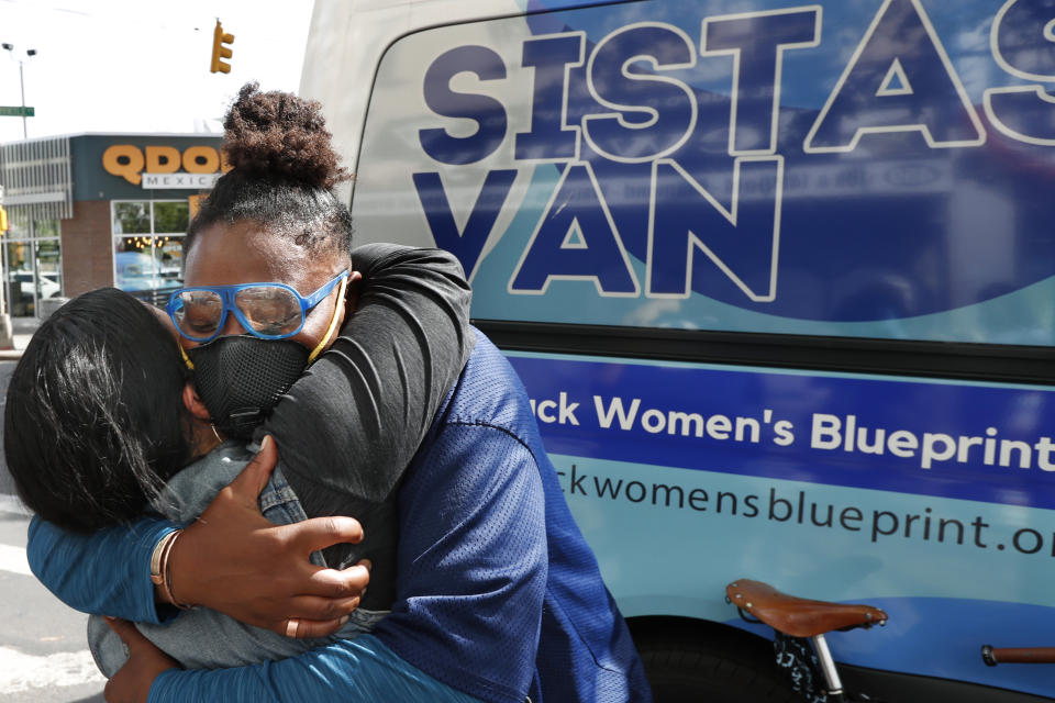 Brooklyn Clayton, right, abandons social distancing precautions to embrace Sistas Van driver Denise Rodriguez after arriving by bicycle to volunteer with the van's crew in handing out free toiletries, personal hygiene products and other items to members of the community in need amid the coronavirus outbreak, Tuesday, May 19, 2020, in New York. Clayton lost her job as a personal chef during the outbreak and now volunteers with the group. (AP Photo/Kathy Willens)