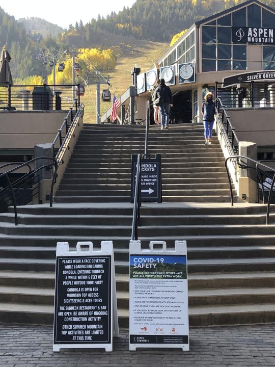 In this Oct. 3, 2020 photo, visitors climb the stairs past COVID-19 safety signs to the gondola at Aspen Mountain in Aspen, Colo. Aspen has become a popular destination during the coronavirus pandemic with its plethora of outdoor activities and precautions being taken by the town. (AP Photo/John Marshall)