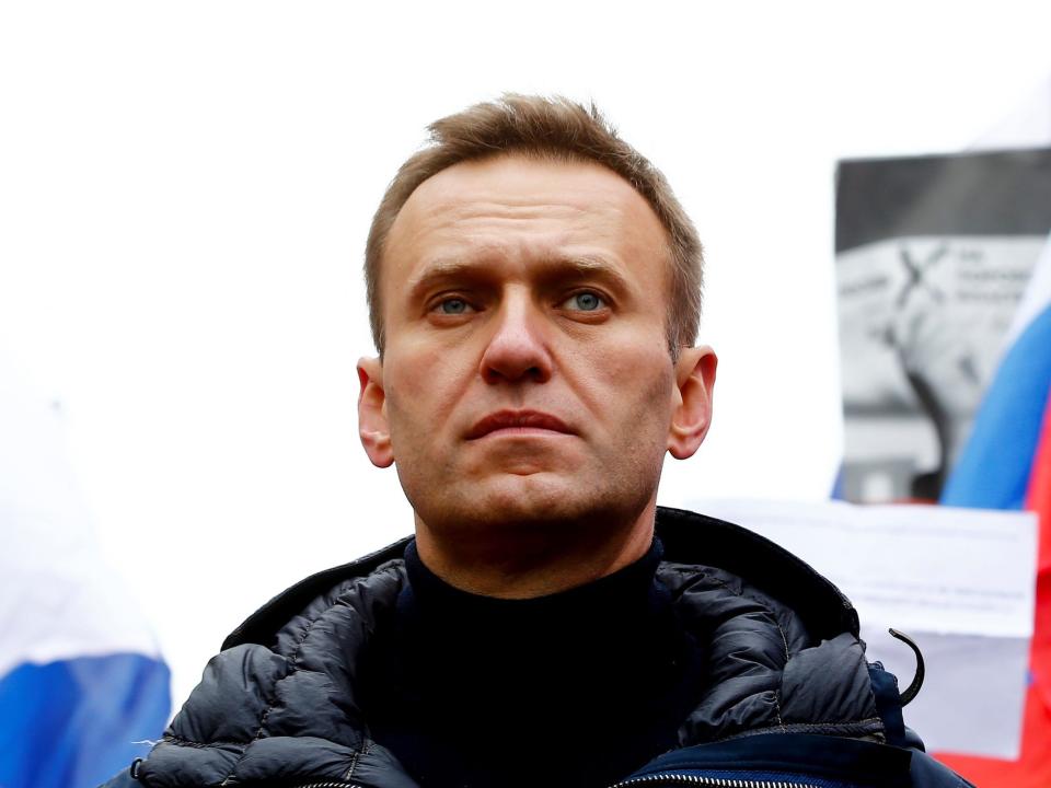 Alexei Navalny wearing a black jacket looks into the distance with Russian flags behind him.