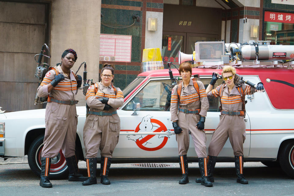 The cast of "Ghostbusters" standing fully suited up in front of their vehicle