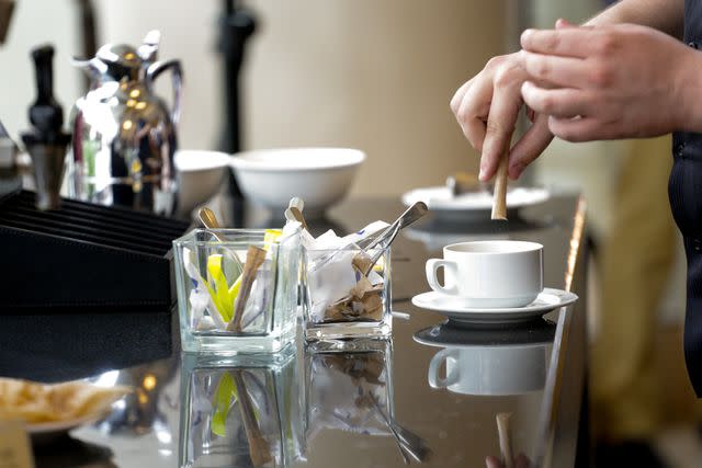 <p>zoranm / Getty Images</p> Hand pouring sweetener from paper packet into coffee cup