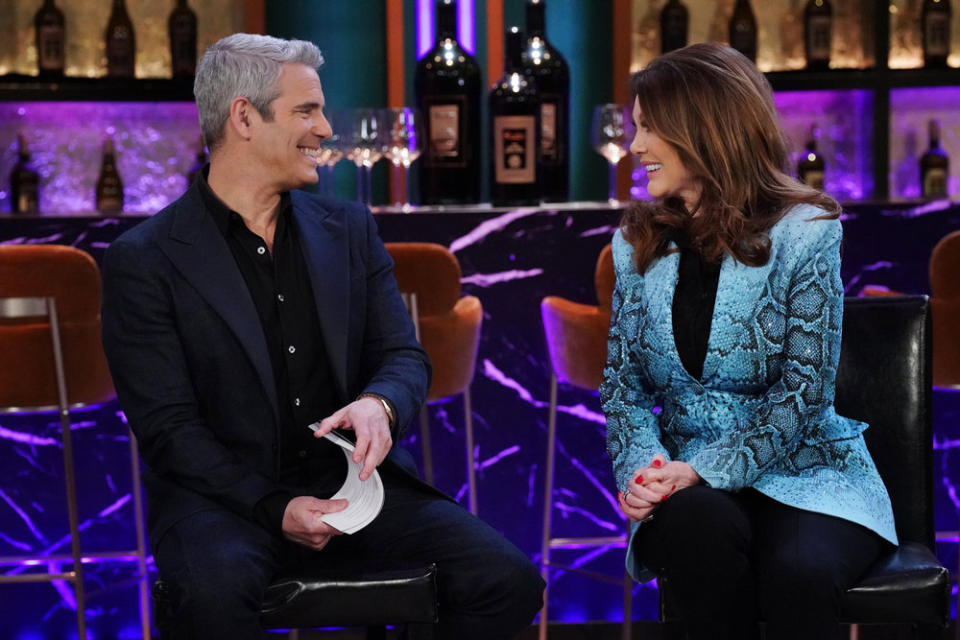 Andy Cohen in a black suit on the left looking at Lisa Vanderpump on the right; she's wearing a blue blazer and they're both smiling at each other