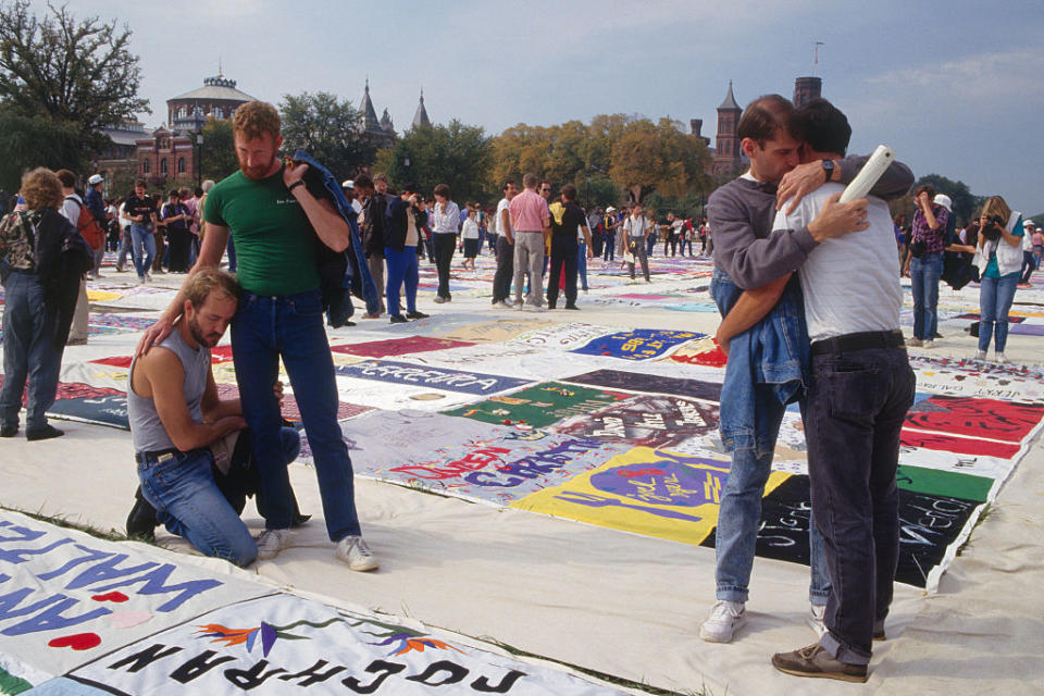 Three individuals among a crowd, on a patchwork of panels with names, embracing in solidarity