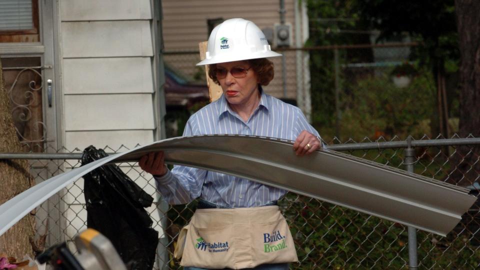 rosalynn carter holds a strip of house siding at a habitat for humanity work site, she is wearing a white hard hat, pink tinted sunglasses, a blue collard shirt with white strips, blue jeans and a fabric tool belt, in the left lower corner of the frame is a circular saw sitting on top of a work bench