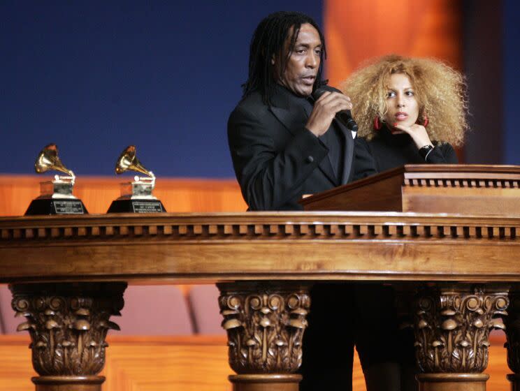 A man in black speaks into a microphone while standing next to his wife and behind a wooden lectern