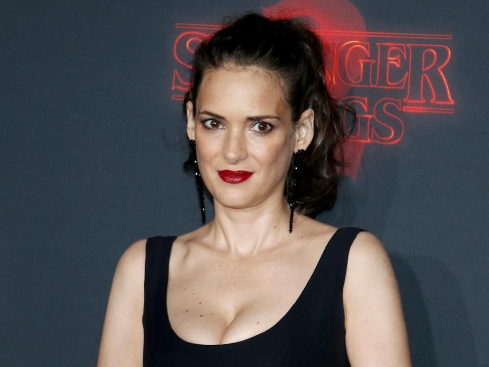 Winona posing with her hair up and in a black tank-top dress in front of a backdrop.