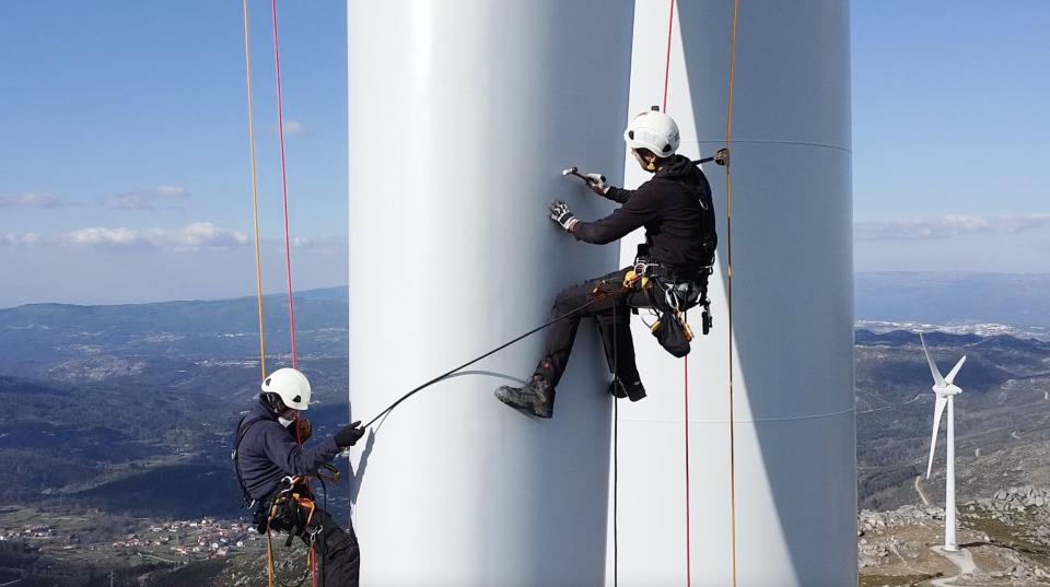 Sardo taps the blade of a wind turbine with a hammer in the mountains of Portugal with his partner close by..